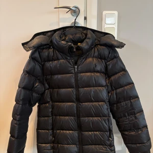 Svart dunjacka från Polo Ralph Lauren - Svart dunjacka från Polo Ralph Lauren med huva och quiltad design. Jackan har dragkedja framtill, två sidofickor och det klassiska Polo-logot på bröstet. 