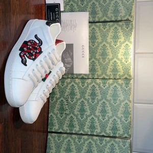 Gucci Ace sneakers med orm - Gucci Ace sneakers i vitt skinn med klassiska gröna och röda ränder på sidan och en broderad orm i rött, svart och vitt. Baksidan har metallicdetaljer i rött och grönt ormskinnsmönster. Snygg och ikonisk modell med snörning och platt sula.