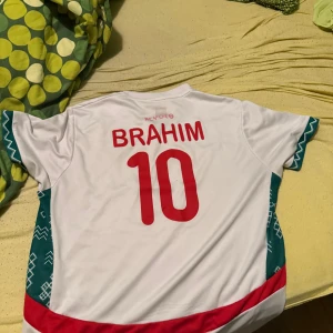 Vit fotbollströja Brahim 10 - Säljer en vit fotbollströja från Kelme med röd text BRAHIM och nummer 10 på ryggen. Tröjan har korta ärmar med gröna och vita grafiska detaljer på sidorna och ärmarna samt en röd sektion längst ner. Perfekt för dig som gillar snygga matchtröjor.