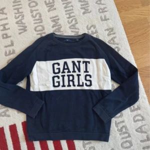 Mörkblå GANT tröja med vit panel - Snygg mörkblå tröja från GANT med vit panel och texten 'GANT GIRLS' framtill. Tröjan har rund halsringning med randig detalj och långa ärmar. Tillverkad i mjuk bomull, perfekt för en chill och stilren look.