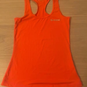 Orange linnetopp från SOC - Säljer en sportig orange linnetopp från SOC i storlek 34. Toppen har brottarrygg och är tillverkad i ett mjukt och stretchigt material som passar perfekt för träning. Enkel och stilren design med liten logga framtill.