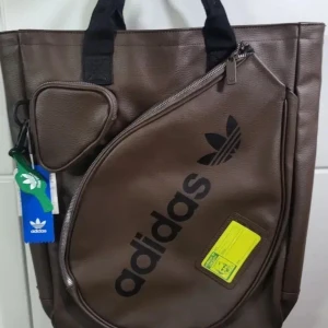 Brun Adidas tennisväska,Helt ny  - Snygg brun axelväska från Adidas med stort svart logotryck och coolt rundat fack med dragkedja framtill. Väskan har svarta handtag och en liten rund ficka på sidan. Perfekt för att bära med sig grejer till träningen . Helt ny och prislappen kvar . Annons gäller bara väska och inget annat.  