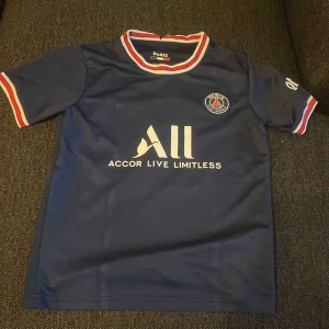 PSG Messi 30 matchtröja - Säljer en officiell PSG fotbollströja med Messi och nummer 30 på ryggen. Tröjan är marinblå med vita tryck, har korta ärmar och detaljer i rött och vitt på krage och ärmslut. Materialet är lätt och andas, perfekt för träning eller match.