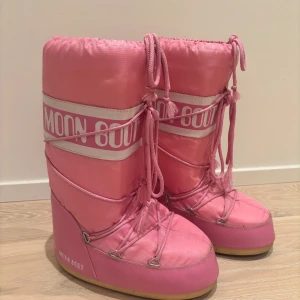 Rosa Moon Boots med snörning - Säljer ett par ikoniska rosa Moon Boots med vit logga och snörning framtill. De har en rund tå, platt sula och är tillverkade i syntetmaterial med vadderad insida. Perfekta för vintern och ger en cool retrostil till din outfit. Pris kan alltid diskuteras!💗storlek 39-41