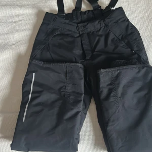 Svarta täckbyxor med hängslen Everest strl.164 cl - Svarta täckbyxor med justerbara hängslen och reflexdetalj på benet. Byxorna har praktiska fickor och elastiska benslut med knapp. Perfekta för vintern och utomhusaktiviteter. Materialet är slitstark syntet som står emot kyla och väta. Använda men klarar fler säsonger! Djur och rökfritt hem. 