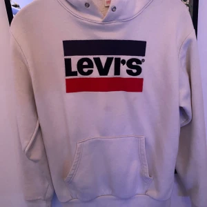 Vit Levi's hoodie med logga - Vit hoodie från Levi's i storlek S med stor, klassisk logga i svart, marinblått och rött på bröstet. Tröjan har huva med snörning, magficka och ribbade muddar. Perfekt för chill dagar och streetstyle. Materialet är mjuk bomull och polyester. De finns dock nåra synliga fläkar, vet inte exakt om den på slide 4 kan gå bort men dom fläkar a mitt på tröjan går inte att få bort