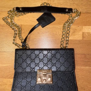 Svart Gucci axelväska med kedja - Snygg svart axelväska från Gucci i skinn med präglat GG-mönster över hela väskan. Väskan har en guldfärgad kedja som axelrem och ett exklusivt lås framtill med Gucci-logga. Perfekt storlek för det viktigaste och har en ficka på baksidan.