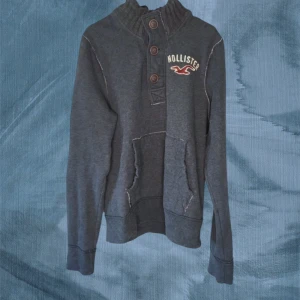 Mörkgrå Hollister hoodie med knappar - Mörkgrå hoodie från Hollister med hög ribbad krage och tre stora knappar framtill. Tröjan har en magficka och broderad logga på bröstet. Långärmad och i mjukt bomullsmaterial, perfekt för kyliga dagar.