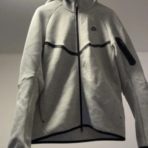Grå Nike zip hoodiejacka - Säljer en grå hoodiejacka från Nike med hel dragkedja och svarta detaljer längs bröstet och ärmen. Jackan har lång ärm, huva och en sportig look som passar perfekt till chill eller träning. Nike-logga på bröstet och ficka på ärmen. Pris ej hugget i sten skriv ifall du vill ha fler bilder, ÄKTA ENDAST SERIÖSA