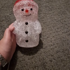 LED Snögubbe Dekorationslampa - Charmig LED-snögubbe med timerfunktion, perfekt som juldekoration. Tillverkad i transparent plast med svart och röd detalj, CE-märkt och IP44-klassad. Fungerar med timer (6h) eller konstant belysning. Inga synliga defekter, fint skick.