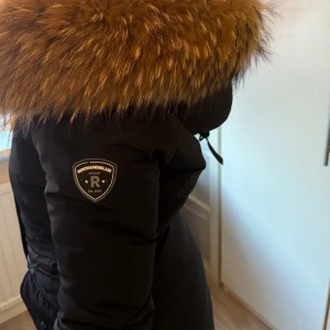 Rockandblue svart dunjacka med päls - Säljer en svart dunjacka från Rockandblue med stor fluffig brun päls runt huvan. Jackan har lång passform, snygga detaljer som logotyp på ärmen och dragsko i midjan. Perfekt för kalla dagar och riktigt stilren look.