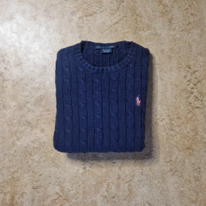 Marinblå kabelstickad tröja Ralph Lauren - Snygg marinblå kabelstickad tröja från Ralph Lauren i 100% bomull. Klassisk rund hals och långärmad modell med röd broderad logga på bröstet. Perfekt för dig som gillar stilrena och tidlösa plagg.