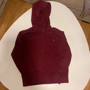 Vinröd hoodie från Ralph Lauren - Säljer en vinröd hoodie från Polo Ralph Lauren med dragkedja och klassisk logga på bröstet. Tröjan har huva, långa ärmar och en stor ficka framtill. Perfekt för chill dagar och enkel att matcha med jeans. Tröjan är för liten och säljs därför