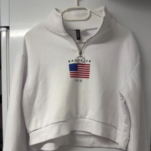 Vit croppad hoodie från H&M Divided - Säljer en vit croppad hoodie från H&M Divided med halv dragkedja och amerikansk flagga samt texten Brooklyn N.Y.C på bröstet. Tröjan har lång ärm, ribbade muddar och är gjord i mjuk bomull och polyester. Perfekt för dig som gillar streetstyle och snygga detaljer.