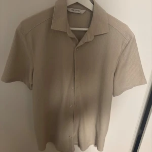 Beige kortärmad skjorta LCW Vision - Stilren beige skjorta från LCW Vision med korta ärmar och klassisk krage. Skjortan har knäppning framtill och är tillverkad i ett lätt, strukturerat tyg som ger en avslappnad vibe. Perfekt för dig som gillar enkel och clean stil.