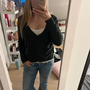 Mörkgrå kofta med v-ringning - Snygg mörkgrå kofta med v-ringad hals och knappar framtill. Modellen är normal i passningen och har långa ärmar samt två fickor framtill. Perfekt att slänga över en topp för en chill look. Materialet känns mjukt och är troligtvis bomull eller en bomullsblandning.