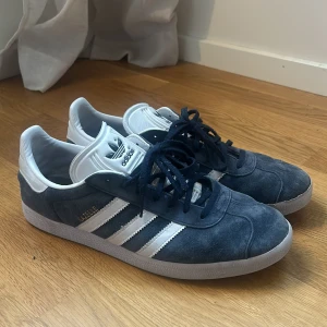 Adidas Gazelle mörkblå sneakers - Säljer ett par klassiska Adidas Gazelle sneakers i mörkblå mocka med vita detaljer. Skorna har andvändts som inne skor i ett år och aldrig haft dem inne. Nypris varierar från 1000-1200kr. Stor sko storlek på : 46 2/3
