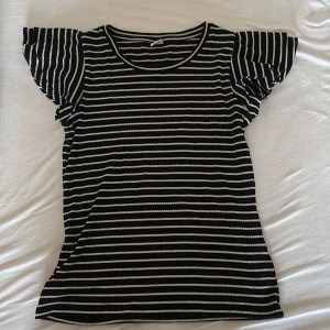 Randig svartvit topp från Lindex - Säljer en svartvit randig topp från Lindex med volangärmar och rund hals. Toppen är ribbad och har en slim passform som sitter snyggt. Perfekt för dig som gillar klassiska mönster och vill ha något som sticker ut med de coola ärmarna.