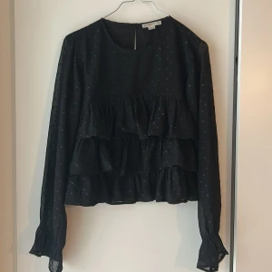 Svart prickig blus med volanger Gina Tricot - Säljer en svart blus från Gina Tricot med glittriga prickar och flera lager volanger framtill. Blusen har långärmade ärmar med lätt rynk vid mudden och rund halsringning. Perfekt för dig som gillar detaljer och en cool look.