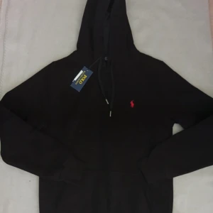 Svart hoodie från Polo Ralph Lauren - Svart hoodie från Polo Ralph Lauren med klassisk röd logga på bröstet. Tröjan har huva med snörning, magficka och långa ärmar. Tillverkad i mjuk bomull som är skön att ha på sig. Perfekt för dig som gillar stilrena och tidlösa plagg.