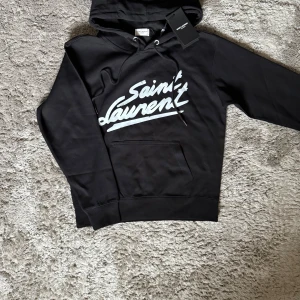 Saint Laurent Hoodie - Exklusiv hoodie från Saint Laurent i svart – en tidlös och lyxig piece med den ikoniska loggan framtill. Perfekt för en stilren och modern look med premiumkänsla.  ♦️ Storlek S – slim passform ⚜️ Helt nytt skick med tagg ♦️ Tillverkad i mjuk bomull av hög kvalitet  📦 Fraktas inom 24 timmar & trygg affär 