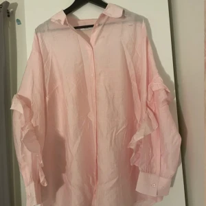 Ljusrosa skjorta med volangärmar - Supersöt ljusrosa skjorta från NA-KD med volangdetaljer längs ärmarna och klassisk krage. Skjortan har knappar framtill och är tillverkad i tunn bomull som ger en luftig känsla. Perfekt för dig som gillar romantiska och trendiga plagg.