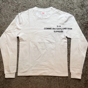 Supreme x Comme des Garçons longsleeve  - Vit långärmad tröja från Supreme x Comme des Garçons med svart tryck på bröstet. Tröjan har rund hals och är gjord i mjuk bomull. Snygg och clean design med texten 'COMME des GARÇONS SHIRT SUPREME' framtill. Perfekt för dig som gillar streetwear.