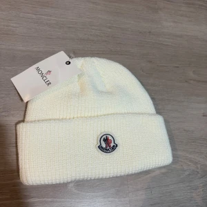 Moncler mössa  - Snygg vit ribbstickad mössa från Moncler med klassisk uppvikt kant och broderad logotyp framtill. Tillverkad i mjukt material som håller dig varm och ger en clean look. Perfekt accessoar för kyliga dagar och enkel att matcha med olika outfits.