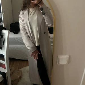 silkes beige trenchcoat  - Snygg beige trenchcoat med klassisk krage och stora svarta knappar. Jackan har ett bälte i midjan och fickor på sidorna. Lång modell som ger en clean och stilren vibe, perfekt för dig som gillar minimalistisk stil.köpt utomlands