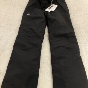 Svarta täckbyxor från 8848 Altitude storlek 150 - Svarta täckbyxor från 8848, storlek 150. Byxorna har rak passform, justerbar midja och slitstarka förstärkningar vid bensluten. Perfekta för vinterns alla äventyr och håller dig både varm och torr. Byxorna är oanvända då de är för små.