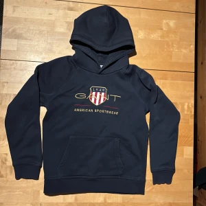Marinblå hoodie från GANT med brodyr - Snygg marinblå hoodie från GANT med broderad logga och sköld på bröstet. Klassisk känguruficka framtill och mjuk insida. Perfekt för dig som gillar sportig och stilren look. Huva och ribbade muddar för extra komfort.