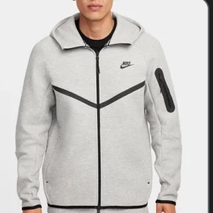 Grå hoodiejacka från Nike - Snygg grå hoodiejacka från Nike med svarta detaljer och dragkedja framtill. Tröjan har huva, lång ärm och en cool svart dragkedjeficka på ena ärmen. Perfekt för en sportig och avslappnad stil.