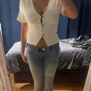 Vit croppad topp från Lager 157 - Säljer en vit croppad topp från Lager 157 i modellen CISSI. Toppen har korta ärmar, v-ringning och snygga knappar framtill. Enkel och stilren design som passar perfekt till jeans eller kjol. Mjuk och stretchig i materialet för en bekväm känsla. Finns även en extra knapp! Tröjan är knappt använd.