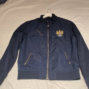 Morris blå bomberjacka med brodyr - Snygg mörkblå bomberjacka från Morris med broderat emblem på bröstet och Union Jack-flagga på insidan. Jackan har flera dragkedjefickor, ribbade muddar och klassisk passform. Perfekt för dig som gillar stilren brittisk vibe.