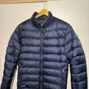 Mörkblå dunjacka från Gant - Snygg mörkblå dunjacka från Gant med quiltad design och dragkedja framtill. Jackan har hög krage, . Perfekt för kyliga dagar och enkel att matcha med det mesta. Köpte från Boozt för 2900.