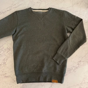 Grå sweatshirt från Solid, S - Mjuk och enkel grå sweatshirt från Solid med rund hals och ribbade muddar. Snygg detalj med broderad logga på ärmen och läderpatch vid nederkanten. Perfekt för chill dagar och passar till jeans eller joggers. Perfekt skick. 