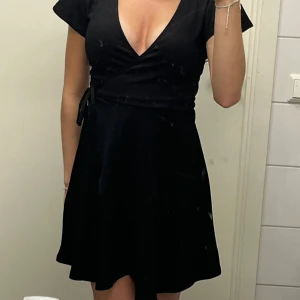 Svart omlottklänning med v-ringning - Supersnygg svart kort omlottklänning med djup v-ringning och knyt i midjan. Klänningen har korta, fladdriga ärmar och en lätt utställd kjol som ger en feminin siluett. Perfekt för dig som gillar en enkel men chic look.