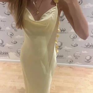 Ljusgul långklänning med blommiga axelband - Superfin ljusgul långklänning från Ginatricot med smala axelband dekorerade med blommor. Klänningen har en mjuk satinliknande yta, draperad urringning och figurnära passform. Perfekt för dig som vill ha en romantisk och trendig look. Helt oanvänd med lappen på