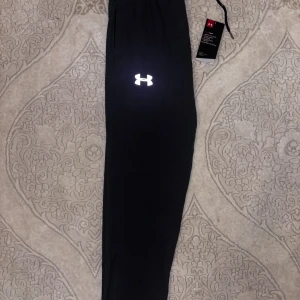 Under Armour träningsbyxor - Snygga gråa träningsbyxor från Under Armour med reflekterande logga. Byxorna är helt nya och taggarna sitter kvar, har dragkedjeförsedda fickor och är gjorda i ett lätt syntetmaterial som andas. Perfekta för gymmet eller löprundan.     