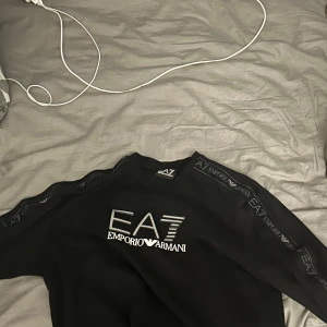 Svart EA7 Emporio Armani sweatshirt - Svart sweatshirt från EA7 Emporio Armani med logga i vitt och silver på bröstet samt tryck längs ärmarna. Tröjan har rund hals och lång ärm, perfekt för dig som gillar stilrena märkeskläder med sportig vibe.
