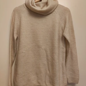 Beige stickad polotröja - Mysig beige stickad polotröja med lång ärm och bred krage. Tröjan har en enkel, klassisk design med ribbade muddar vid ärmslut och nederkant. Perfekt för att hålla värmen under kyliga dagar och passar dig som gillar en chill och stilren look.