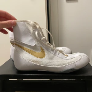 Nike Machomai boxningsskor vit/guld - Nike Machomai boxningsskor i vitt med guldigt swoosh på sidan. Skorna är höga med snörning hela vägen upp och har en lätt och ventilerande ovandel i syntetmaterial. Yttersulan är grå och ger bra grepp. Perfekta för dig som vill ha stilrena och sportiga boxningsskor.