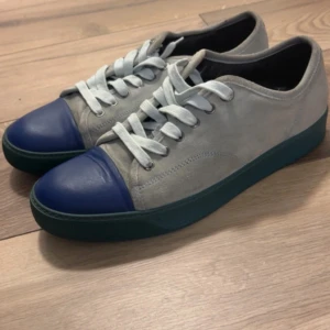 Lanvin sneakers i mocka och blått skinn - Snygga sneakers från Lanvin i ljusgrå mocka med blått skinn över tån och gröntonad sula. Skorna har vita snören och en klassisk låg profil. Perfekta för dig som gillar stilrena men ändå unika detaljer.