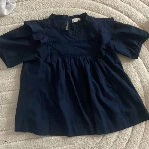 Mörkblå blus med volang från Gina Tricot - Snygg mörkblå blus från Gina Tricot i storlek 158/164. Blusen har korta ärmar, volangdetaljer vid axlarna och en liten öppning med knapp i nacken. Tillverkad i mjukt och lätt bomullsmaterial, perfekt för dig som gillar stilrena och trendiga plagg.