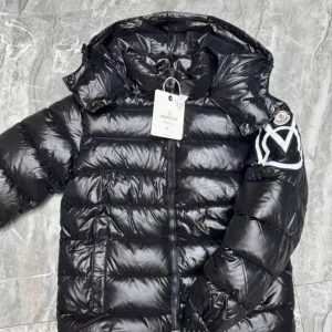 Svart Moncler pufferjacka med huva - Säljer en svart pufferjacka från Moncler med glansig finish och huva. Jackan har dragkedja framtill, två sidofickor och en stor vit logga på ena ärmen. Perfekt för kalla dagar och har en riktigt snygg streetstil.