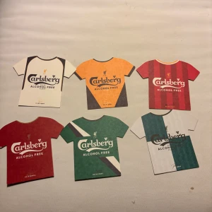 Liverpool Carlsberg retro fotbollströjor - Säljer sex stycken retroinspirerade Liverpool FC fotbollströjor i pappersformat med . Tröjorna har olika färger och mönster, med namn och nummer på baksidan som Carragher, Fowler, Rush, Garcia, Hyypiä och Barnes. Perfekt för samlare eller fans!