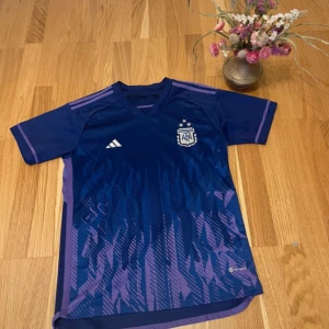 Argentina Adidas blå fotbollströja - Snygg Argentina fotbollströja från Adidas i mörkblått och lila med korta ärmar. Tröjan har AFA-emblem på bröstet, Adidas-logga och ett grafiskt mönster framtill. Tillverkad i lätt och ventilerande polyester, perfekt för match eller träning.