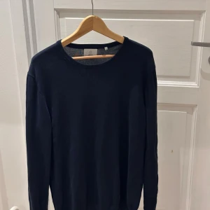 Mörkblå stickad tröja från Bläck - En stilren mörkblå stickad tröja från Bläck i storlek L. Tröjan har rund halsringning, ribbade muddar och långärmad passform. Perfekt för dig som gillar enkel och clean stil. Materialet är mjukt och känns skönt mot huden.