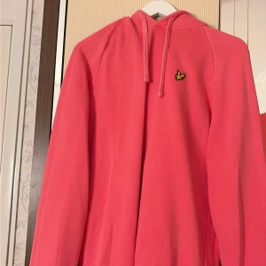 Rosa hoodie från Lyle & Scott - Snygg rosa hoodie från Lyle & Scott med klassisk gul fågel-logga på bröstet. Tröjan har huva med snörning och är tillverkad i mjuk bomull. Perfekt för dig som vill ha en färgglad och bekväm look. Passar dig som gillar streetwear och stilrena detaljer.