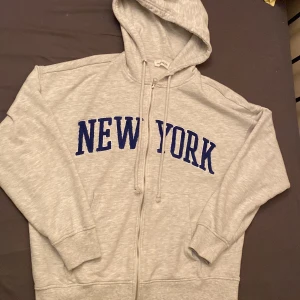 Grå hoodie med blått New York-tryck - Snygg grå hoodie från Even&Odd med blått New York-tryck på bröstet. Tröjan har huva med snörning, dragkedja framtill och två fickor. Perfekt för chill dagar och streetstyle. Mjuk bomullsmix och långärmad modell.
